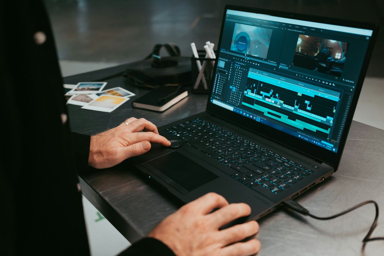 An editor's hands on a laptop keyboard with a video timeline on screen, on a dark desk with printed photos and a camera beside
